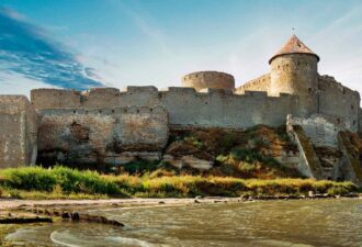 Середньовічний замок на березі річки, оточений зеленими насадженнями та скелястими пагорбами, з величними вежами та кам'яними стінами, що зберігають історію і архітектурну спадщину.