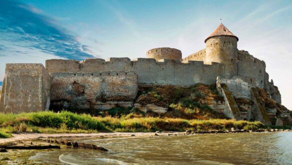 Середньовічний замок на березі річки, оточений зеленими насадженнями та скелястими пагорбами, з величними вежами та кам'яними стінами, що зберігають історію і архітектурну спадщину.