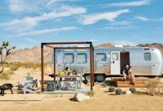 Сім'я відпочиває на природі біля стильного автодому Airstream у пустельному ландшафті, обладнаному зоною для барбекю та затишними меблями. На фоні видно гори та блакитне небо.