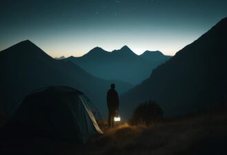 Людина стоїть біля палатки в горах у темряві, тримаючи ліхтарик, який м’яко освітлює навколишню територію; нічне небо темне з кількома видимими зорями, підкреслюючи спокій і самоту дикої природи.