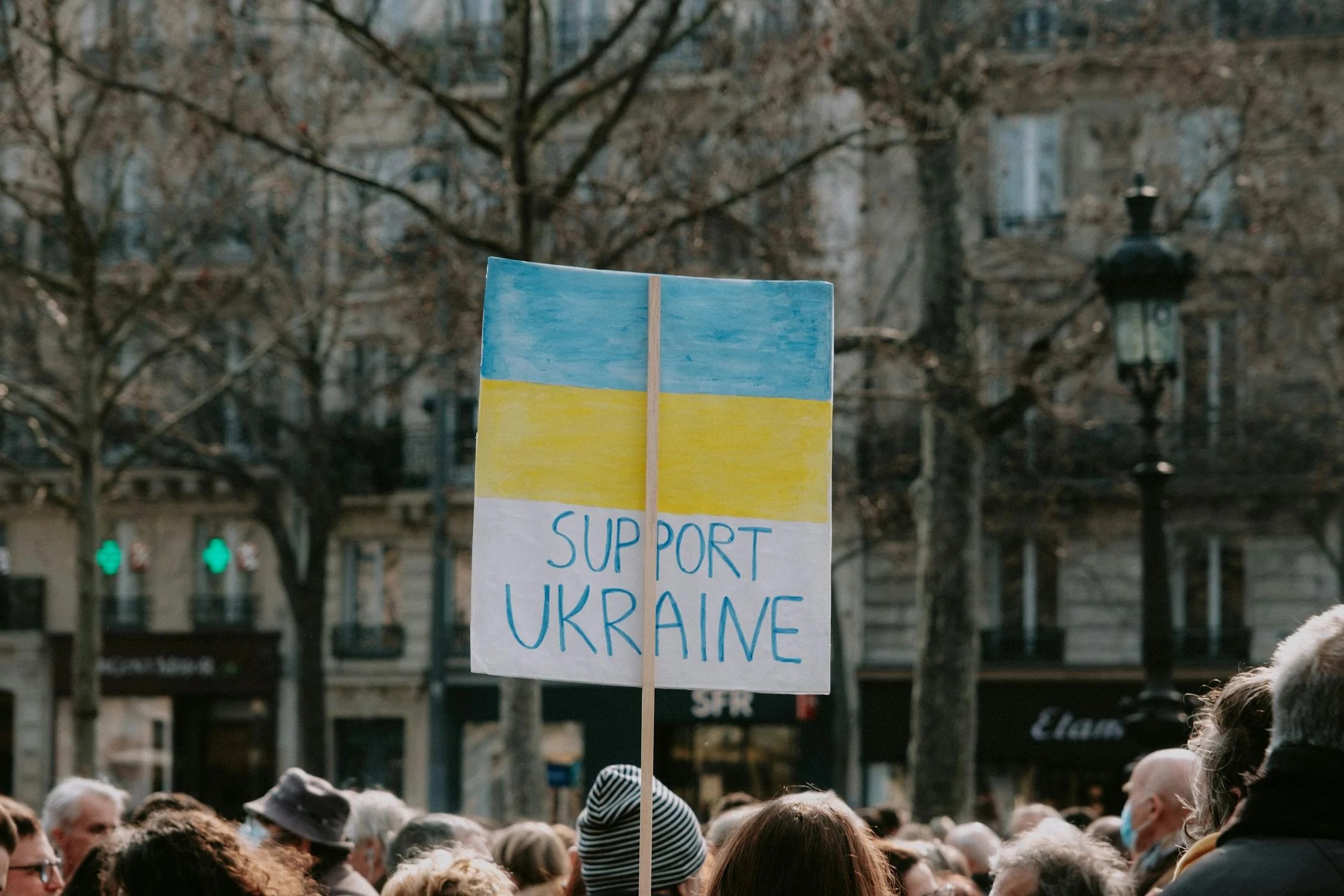 Люди на мітингу тримають плакат з написом на підтримку України, на фоні міського пейзажу.