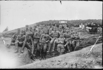Чорно-біла історична фотографія військових часів Першої світової війни. Група солдатів у формі сидить і стоїть перед укріпленою траншеєю, обличчя серйозні, деякі з них мають біноклі та військові нагороди. Позаду видно бліндаж із укріпленим дахом, зробленим з колод і землі. Атмосфера передає дух фронтового життя та військової спільності.