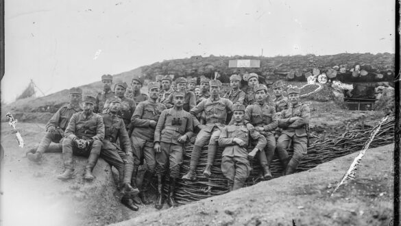 Чорно-біла історична фотографія військових часів Першої світової війни. Група солдатів у формі сидить і стоїть перед укріпленою траншеєю, обличчя серйозні, деякі з них мають біноклі та військові нагороди. Позаду видно бліндаж із укріпленим дахом, зробленим з колод і землі. Атмосфера передає дух фронтового життя та військової спільності.