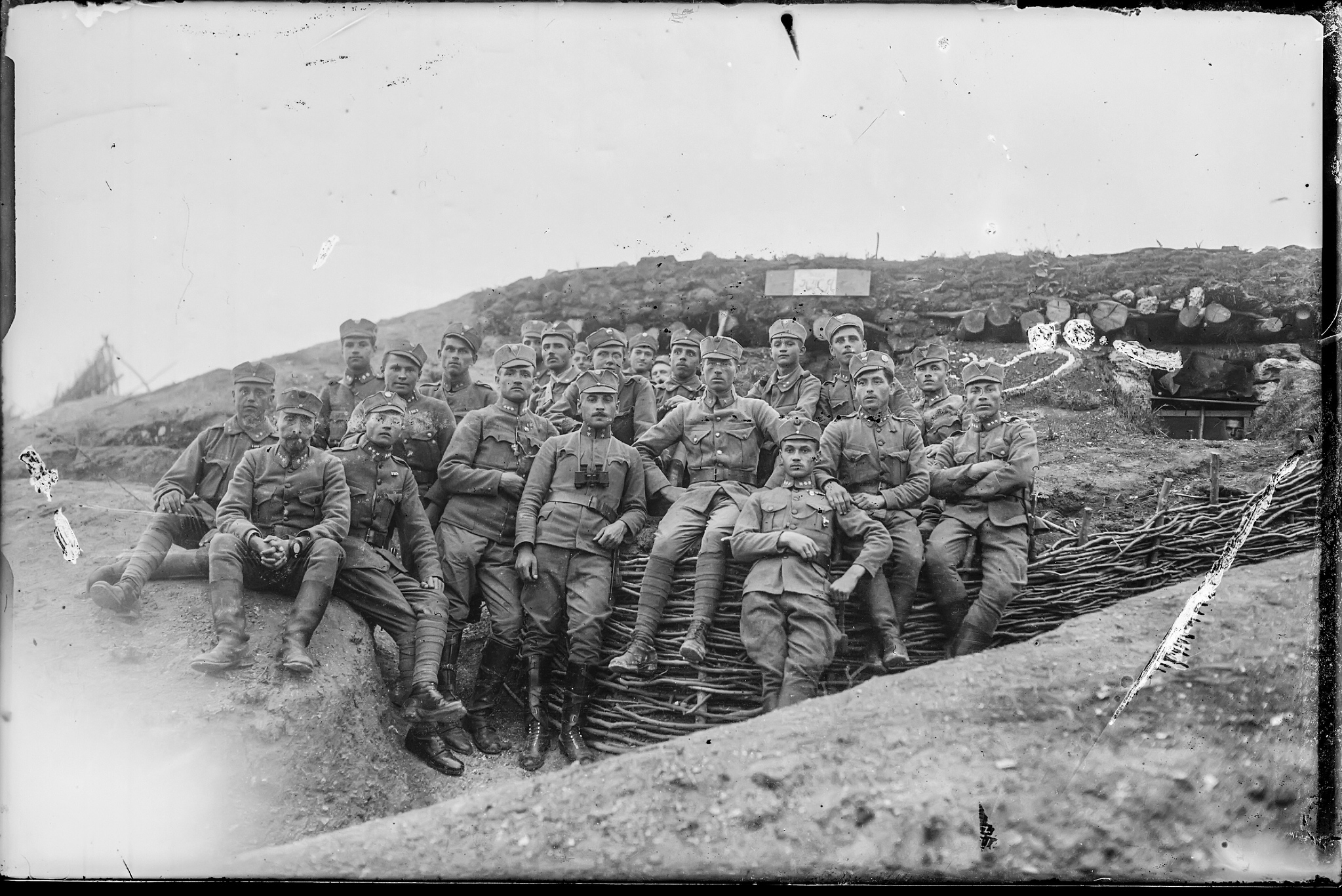Чорно-біла історична фотографія військових часів Першої світової війни. Група солдатів у формі сидить і стоїть перед укріпленою траншеєю, обличчя серйозні, деякі з них мають біноклі та військові нагороди. Позаду видно бліндаж із укріпленим дахом, зробленим з колод і землі. Атмосфера передає дух фронтового життя та військової спільності.