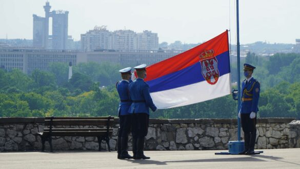 Солдати в формі піднімають національний прапор Сербії на фоні міського пейзажу з зеленими насадженнями.