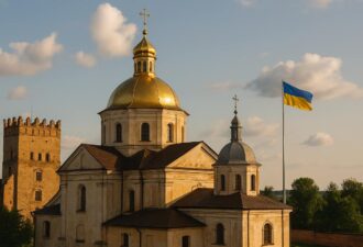 Золотоголова церква поруч із середньовічною вежею та прапором України на тлі вечірнього неба з хмарами.