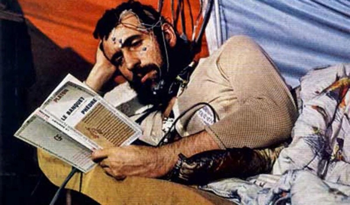 На фото зображено чоловіка, який читає книжку, лежачи на ліжку, підключений до електродів — імовірно, це частина експерименту, пов’язаного зі сном або тривалим перебуванням у замкненому просторі. Схоже, що це фото з радянського експерименту 70-х чи 80-х років, можливо — в рамках досліджень ізоляції або підготовки до космічних польотів. Атмосфера виглядає як імпровізований намет або герметичний простір.