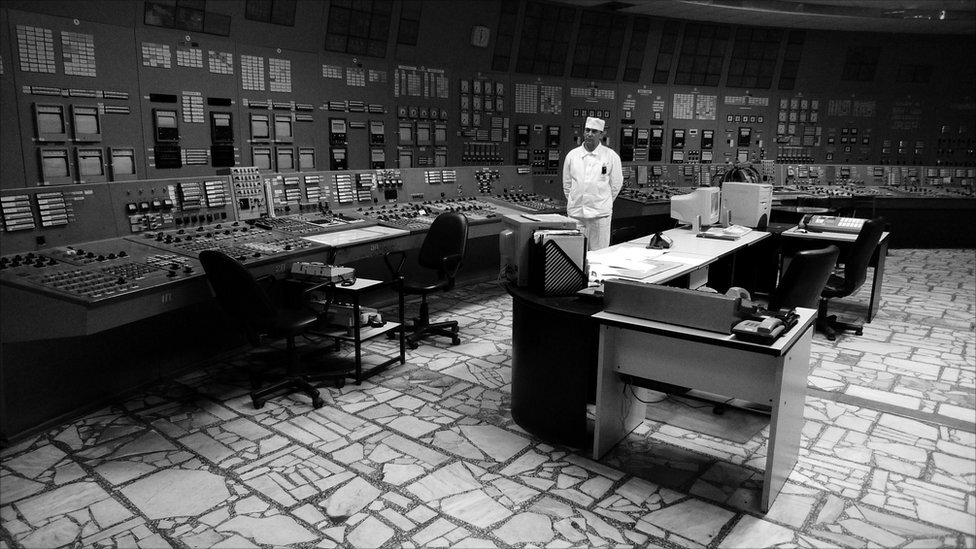 Оператор у білому спецодязі стоїть у диспетчерській залі Чорнобильської АЕС, серед панелей із численними кнопками та моніторами.