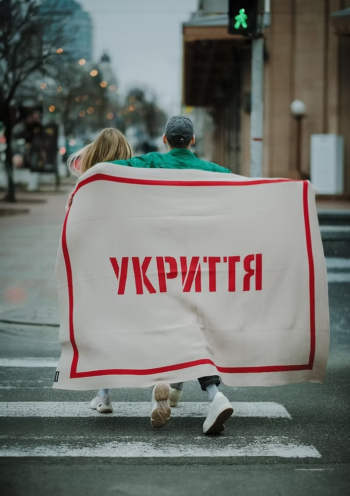 Двоє людей, які переходять пішохідний перехід у місті, тримаючи за плечима плед з написом «УКРИТТЯ» (великими червоними літерами). Плед обрамлений червоною рамкою. У кадрі видно зелене світло світлофора для пішоходів. Атмосфера — міська, рання весна чи осінь