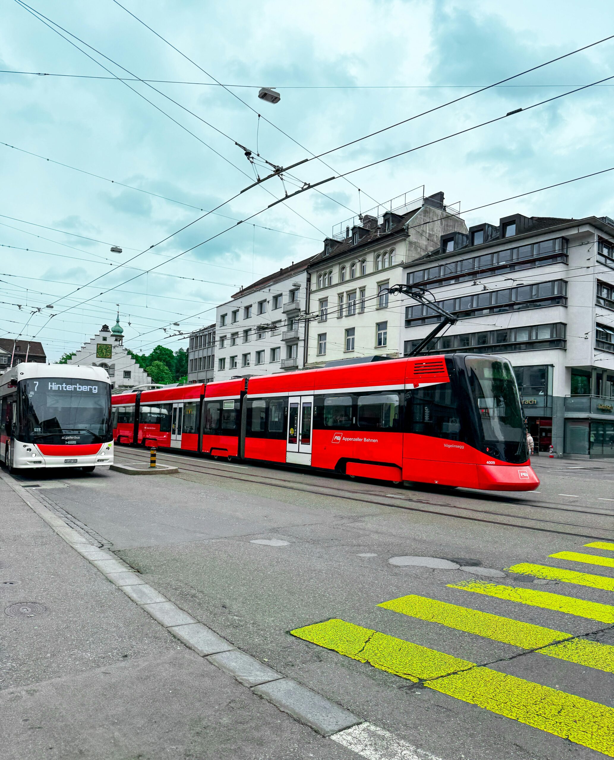 Сучасний червоний трамвай Appenzeller Bahnen і автобус маршруту №7 “Hinterberg” на міській вулиці Швейцарії, з історичними будівлями на задньому плані.
