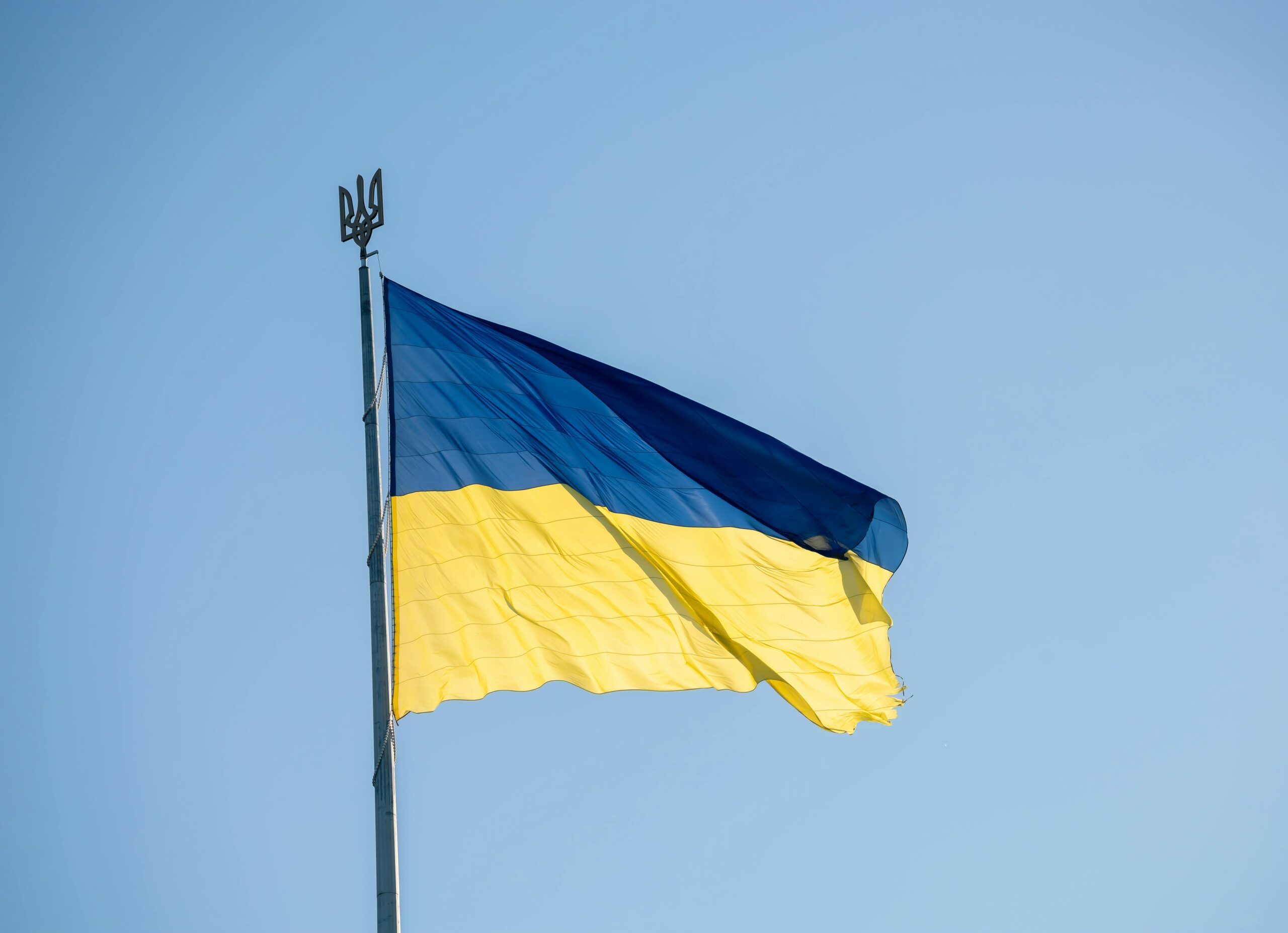 Український прапор майорить на вітрі на тлі чистого блакитного неба, зверху — тризуб на флагштоку.