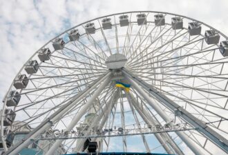 Велике оглядове колесо на Контрактовій площі в Києві з українським прапором посередині конструкції, на фоні хмарного неба.