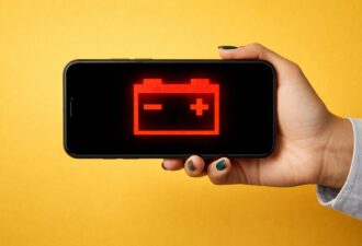 Рука тримає смартфон на жовтому фоні, на екрані телефона чорний фон із червоним значком розрядженої батареї, мінімалістичний горизонтальний кадр.