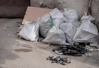 Мішки з будівельним сміттям стоять біля старої бетонної стіни поруч із купою старих телефонів і використаних батарейок, розкиданих на землі. Атмосфера занедбаного міського двору підкреслюється пилом, уламками бетону та приглушеним природним світлом.