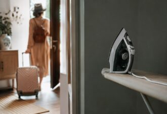Праска стоїть на прасувальній дошці в центрі уваги, тоді як на задньому плані людина з валізою виходить із квартири перед подорожжю. М’яке природне світло створює атмосферу зборів у відпустку, а композиція передає знайоме багатьом хвилювання про те, чи всі електроприлади вимкнені перед від’їздом.